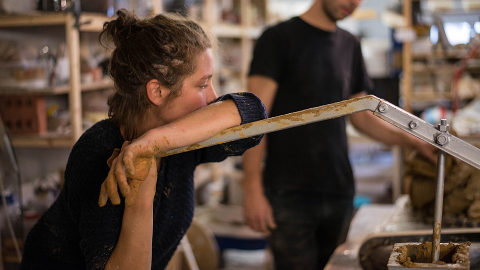 Woman holding a ceramic extruding machine lever in a ceramic workshop with a man in the background 