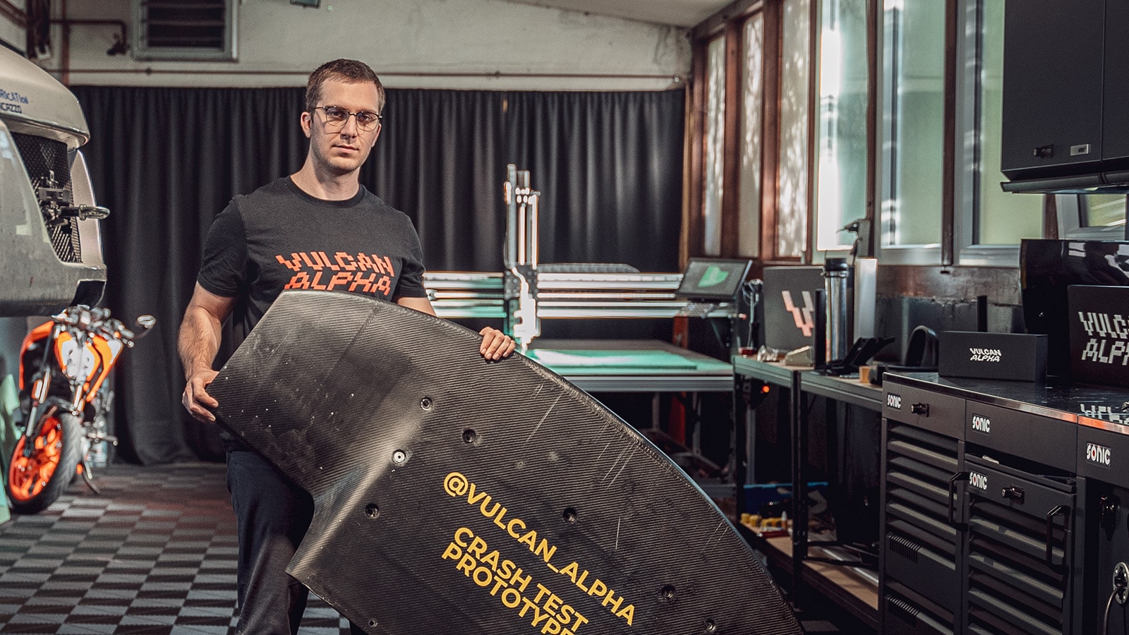 Man holding a crash test prototype of carbon fiber aerodynamic part for racing cars with a CNC milling machine milling foam in the background