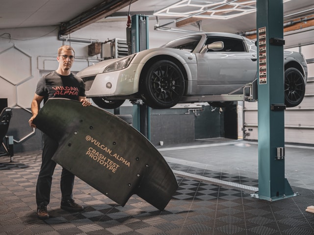 Man holding a crash test prototype of carbon fiber aerodynamic part for racing cars with a car in the background